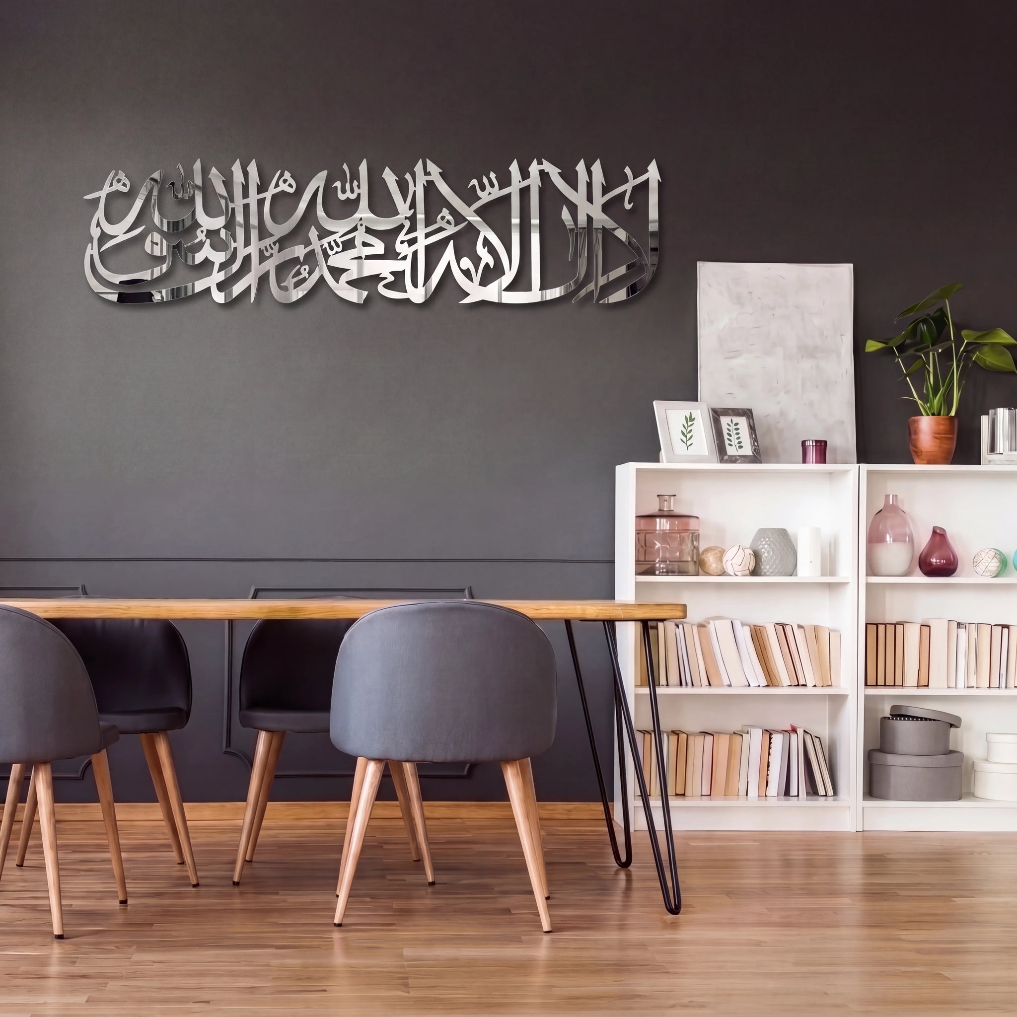 A modern interior scene with a dark gray wall featuring Shahada Kalima large Stainless Steel Arabic calligraphy wall art above a wooden dining table and gray chairs. White bookshelves with books and decor sit to the right, and a potted plant is visible on top.