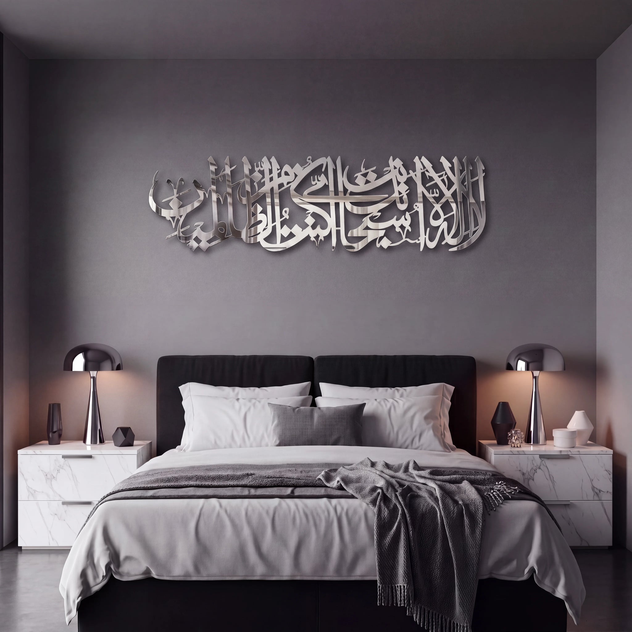 A modern bedroom featuring a large, Ayat Kareema metallic Arabic calligraphy wall art piece mounted on a dark gray wall above a black bed with white and gray bedding. The room is styled symmetrically with white marble nightstands, sleek Chrome silver table lamps, and minimalist decor.