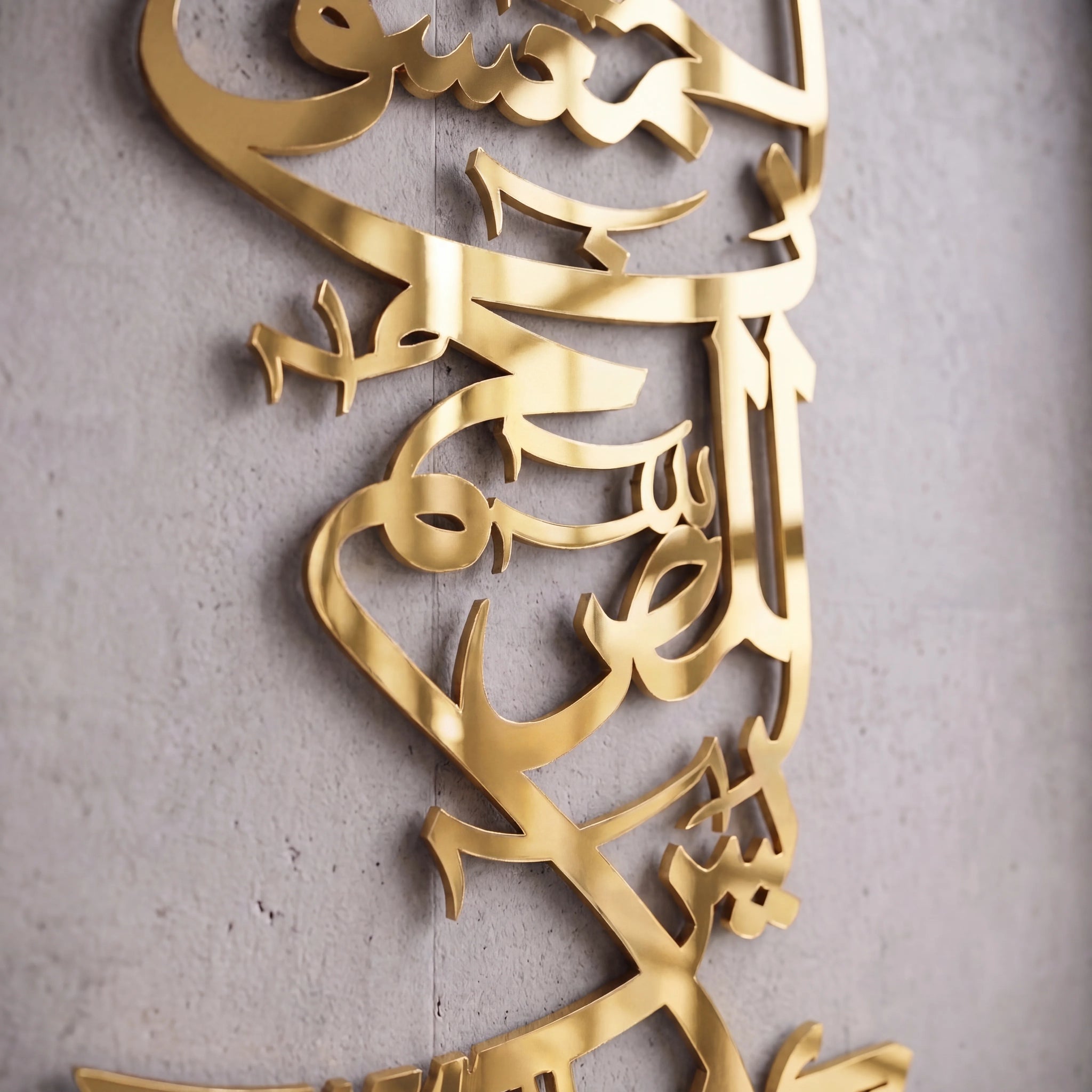 Shiny Gold metallic Islamic calligraphy wall art mounted on a textured gray concrete wall.
