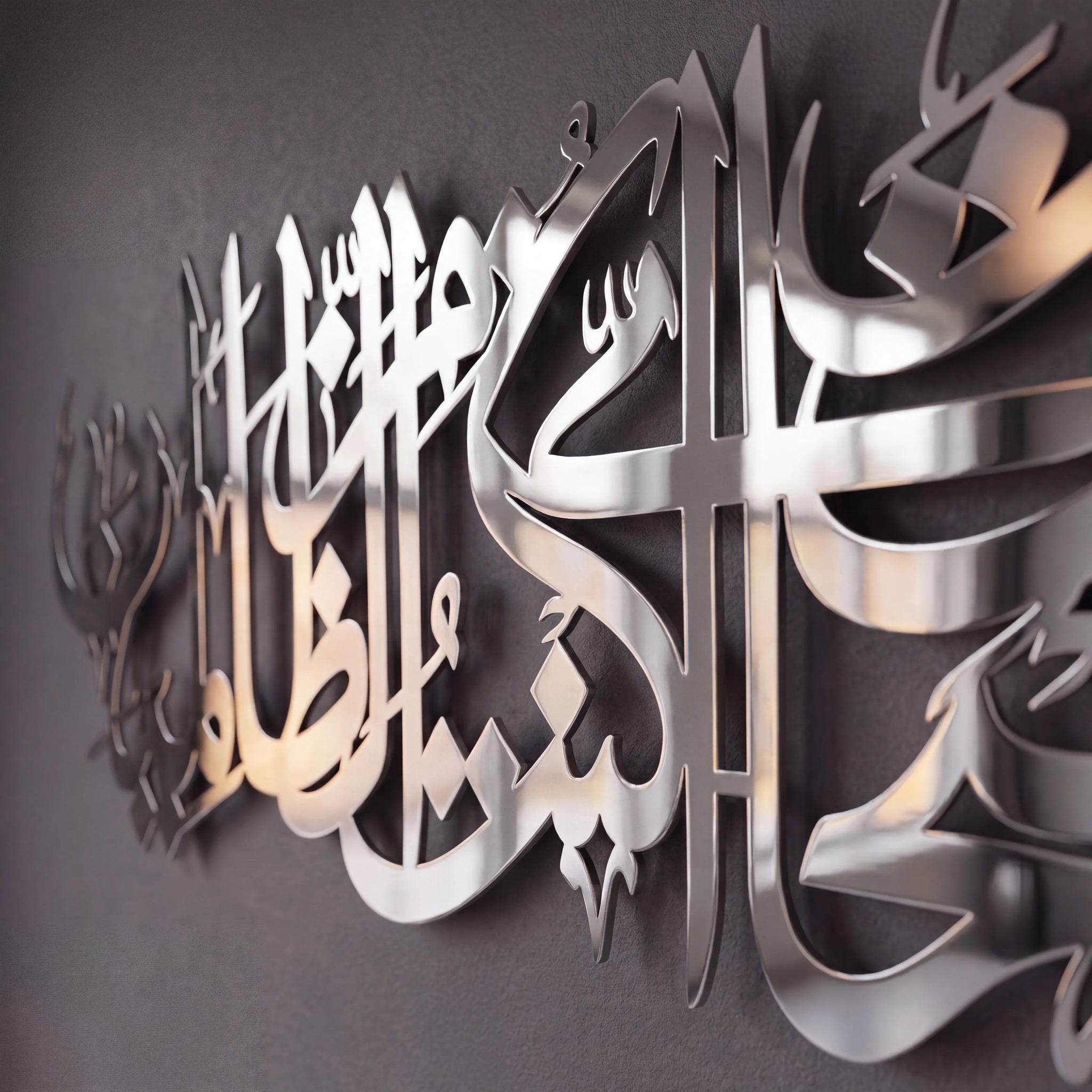 Metallic Islamic wall art featuring Arabic calligraphy in a polished silver finish, mounted on a dark gray textured wall.