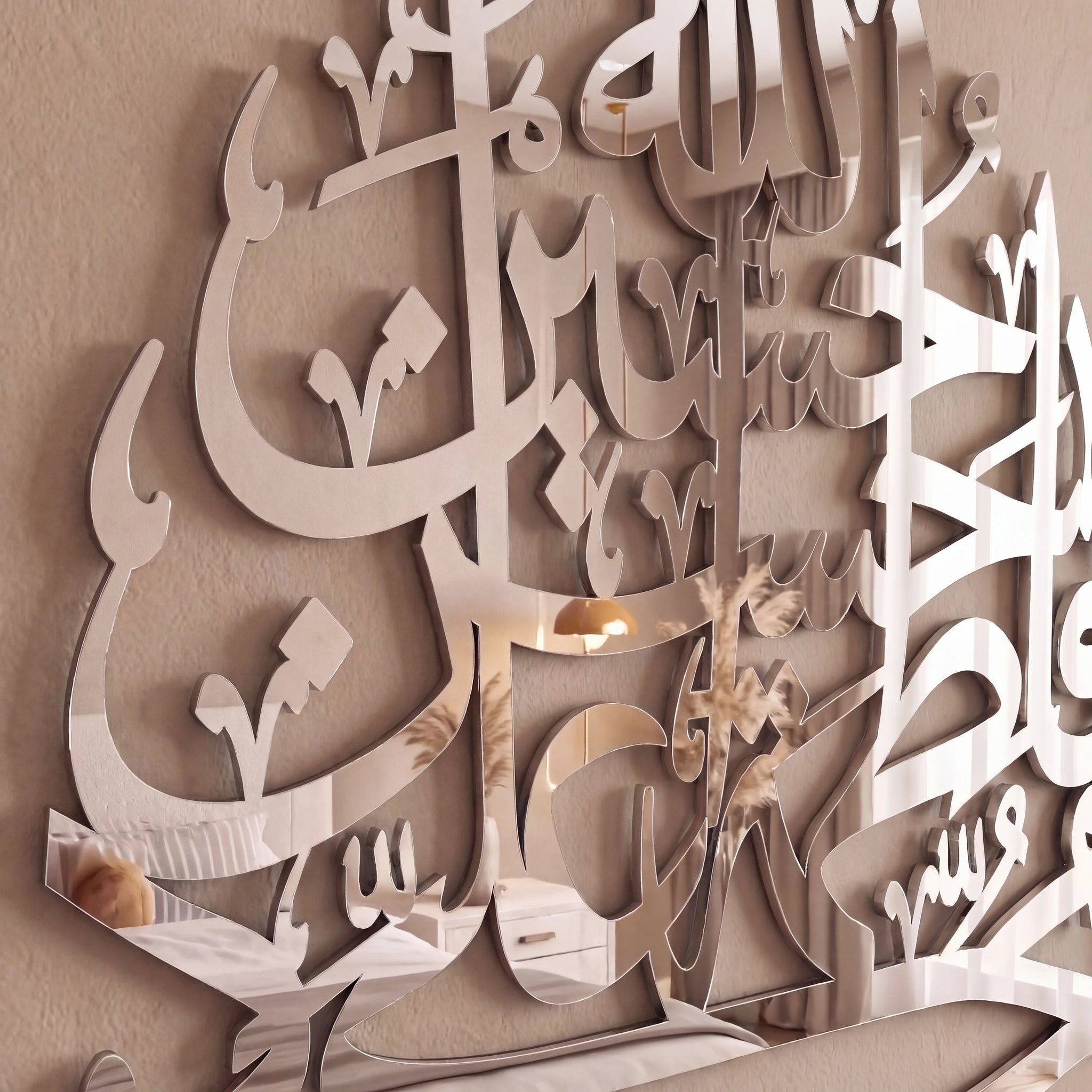 Large reflective chrome silver Arabic calligraphy wall art mounted on a beige wall, with a small warm light reflection visible in the mirrored surface.