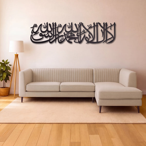 Shahada Kalima Black metal Islamic wall art featuring Arabic calligraphy of 'Allah' and 'Subhan Allah', displayed on a beige wall above a modern beige sectional sofa in a styled living room.
