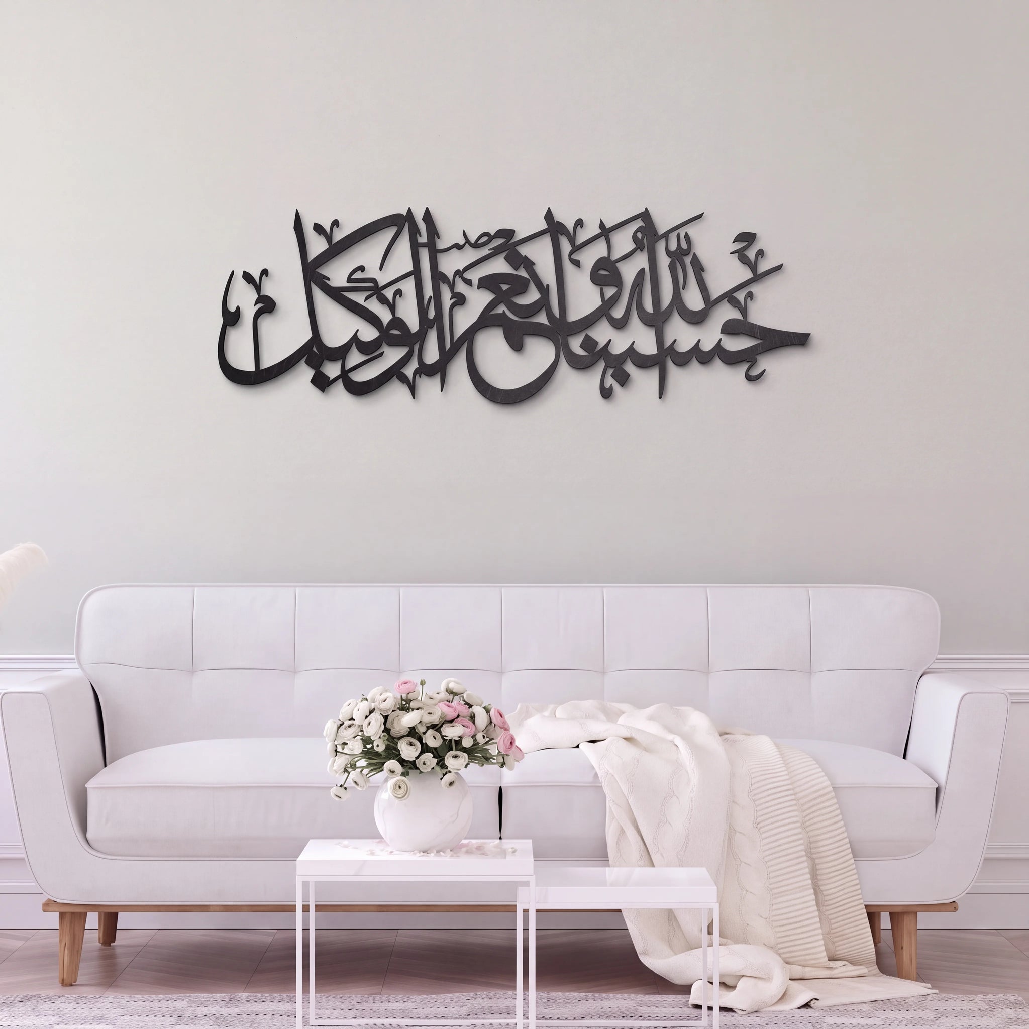 Black metallic Arabic calligraphy wall art mounted on a light gray wall above a modern white sofa in a styled living room.