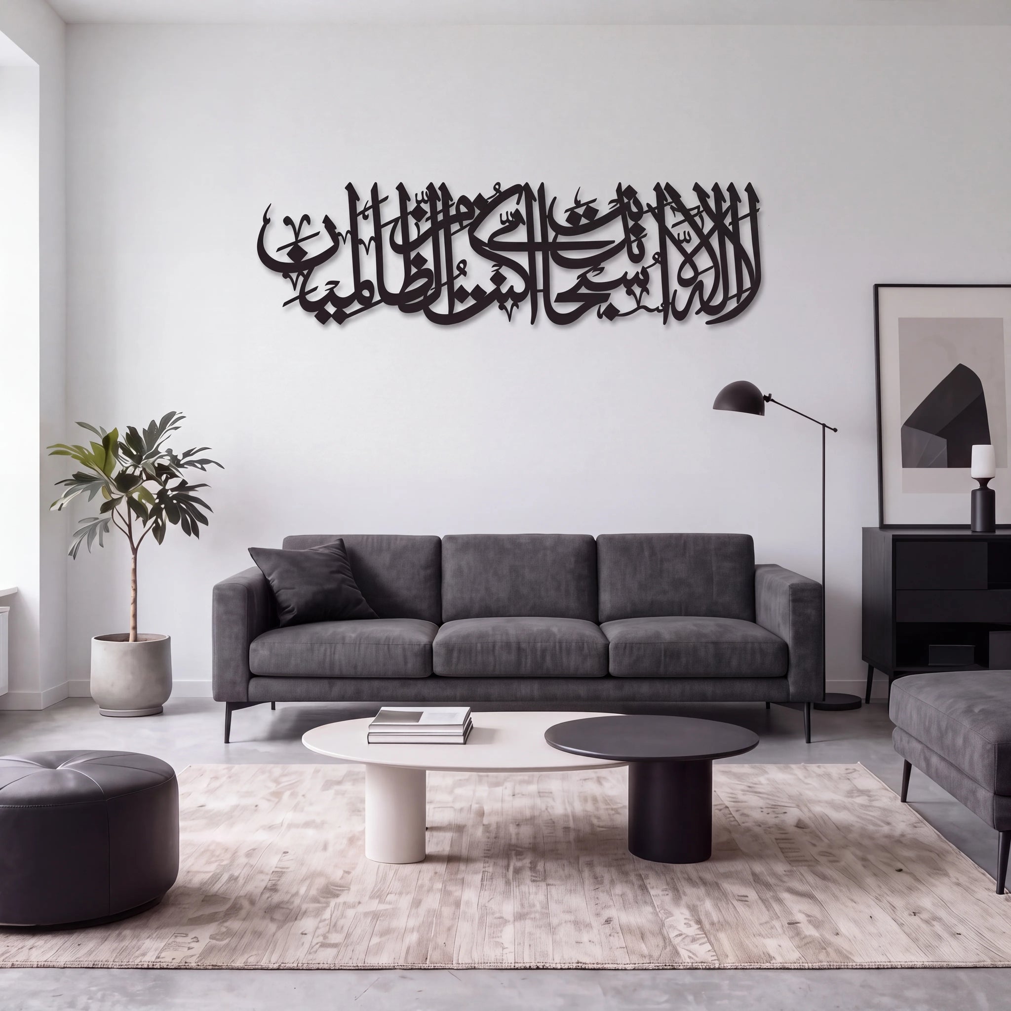 A modern living room featuring a large Ayat Kareema black metal Islamic calligraphy wall art piece mounted on a white wall above a gray fabric sofa. The room is styled with a light-colored rug, a round ottoman, a potted plant, and minimalist furniture.