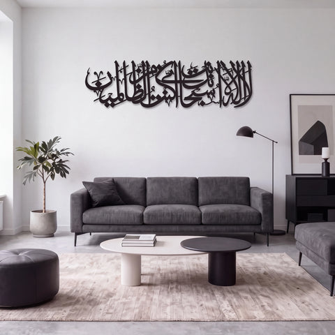 A modern living room featuring a large Ayat Kareema black metal Islamic calligraphy wall art piece mounted on a white wall above a gray fabric sofa. The room is styled with a light-colored rug, a round ottoman, a potted plant, and minimalist furniture.