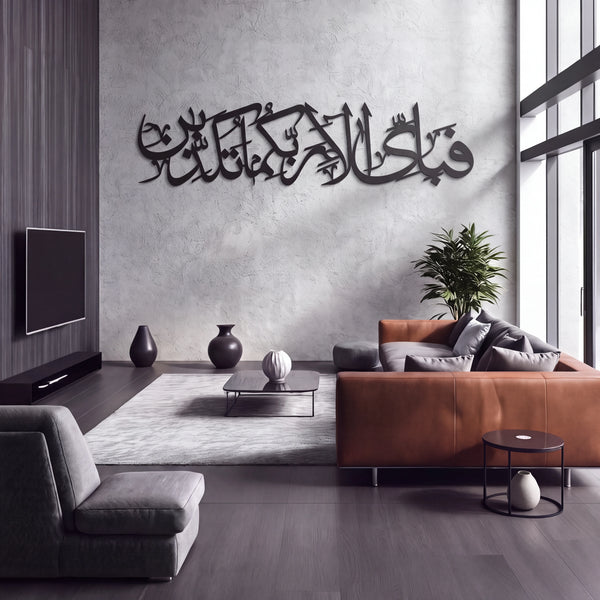 A modern living room interior with a Fabi Ayyi Alai Rabbikuma Tukazziban large black metal Arabic calligraphy wall art piece mounted on a textured gray wall. The room features a brown leather sofa, a gray armchair, a light gray rug, dark wood flooring, and a potted plant.