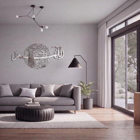 A modern living room with a gray sofa, round black ottoman, white rug, and a large Darood Shareef Arabic calligraphy Chrome Silver wall art piece mounted on the gray wall above the sofa.