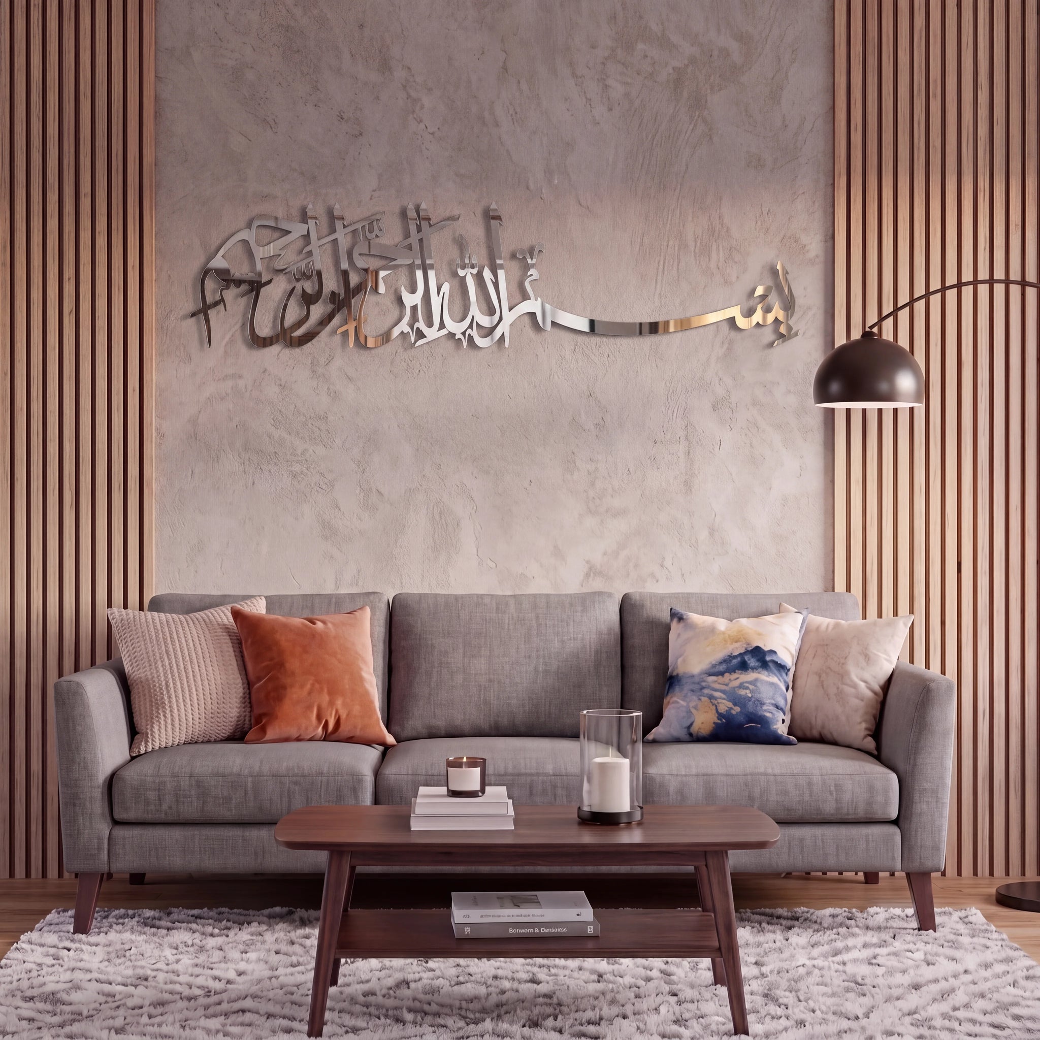 A modern gray fabric sofa in a styled living room setting, decorated with neutral and patterned throw pillows, a dark wood coffee table, a textured rug, and a concrete wall featuring Chrome Silver metallic Bismillah Arabic calligraphy wall art.