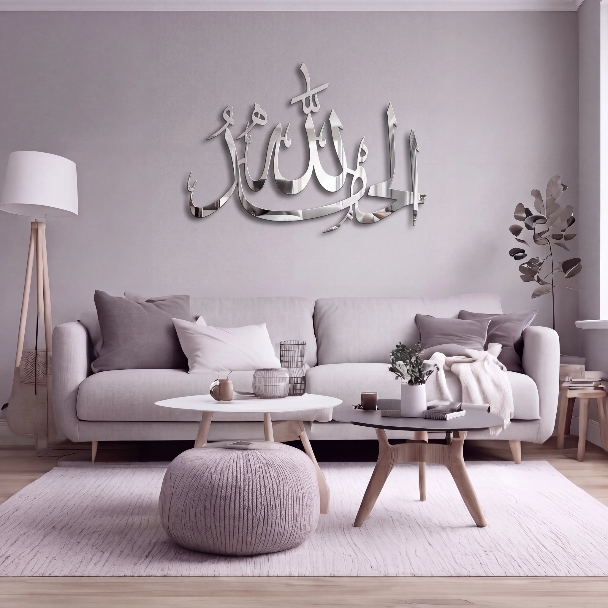 A modern living room with a light gray sofa, minimalist coffee tables, a round pouf, and a large reflective Chrome Silver Alhamdulillah Arabic calligraphy wall art piece mounted on the wall above the couch.