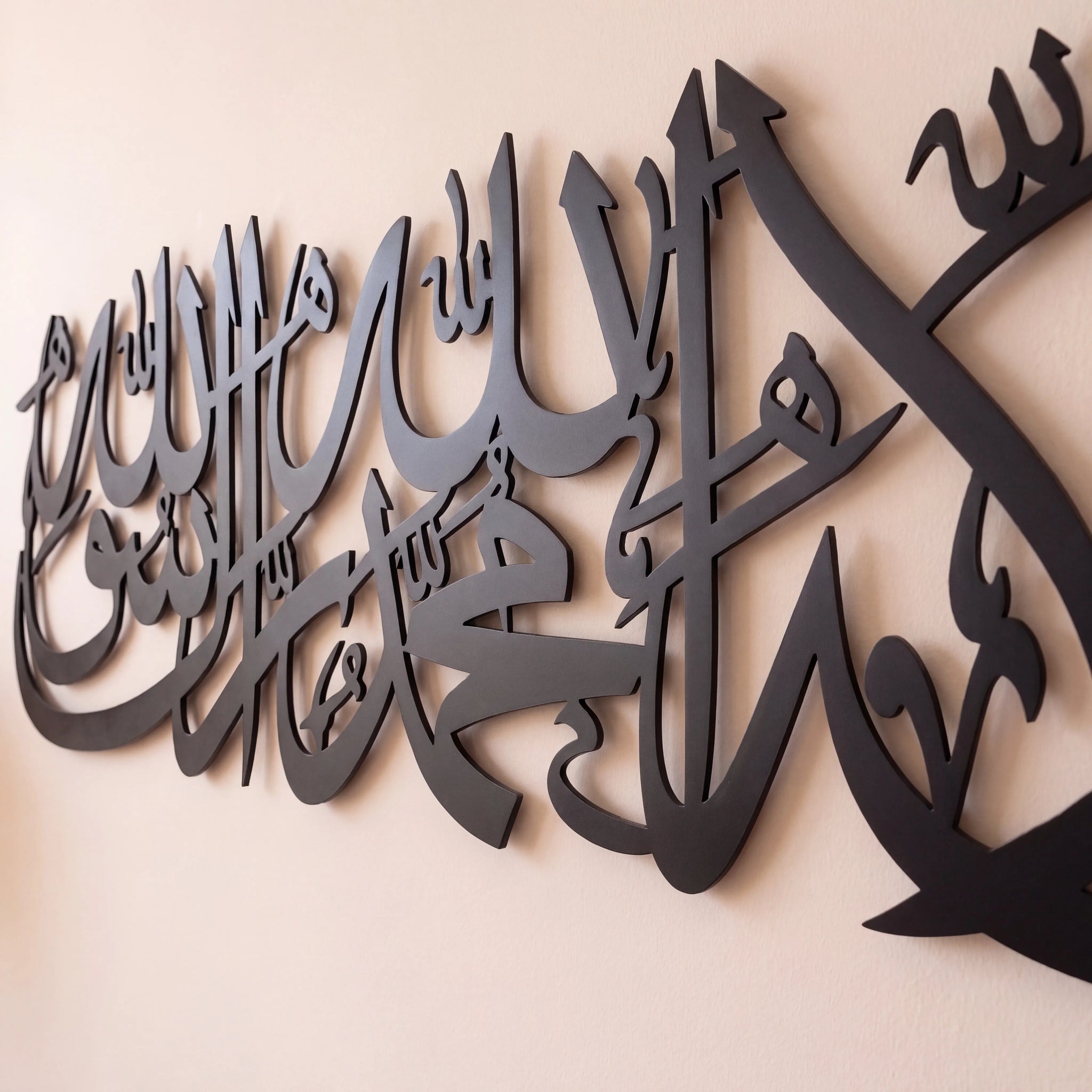 Shahada Kalima Black metal Islamic wall art featuring Arabic calligraphy, mounted on a light beige wall.