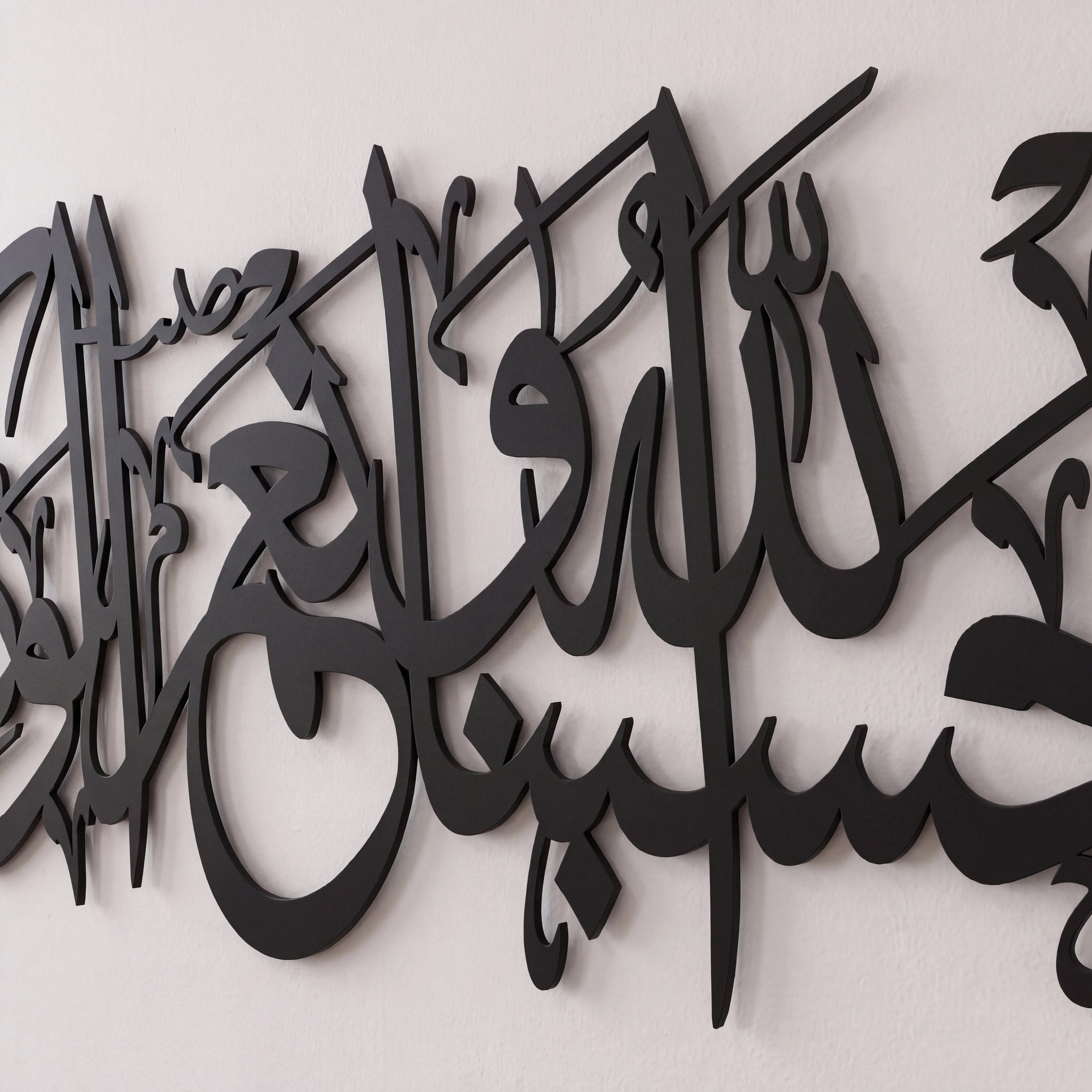Black Quran Ayat metal Arabic calligraphy wall art mounted on a light off-white textured wall.