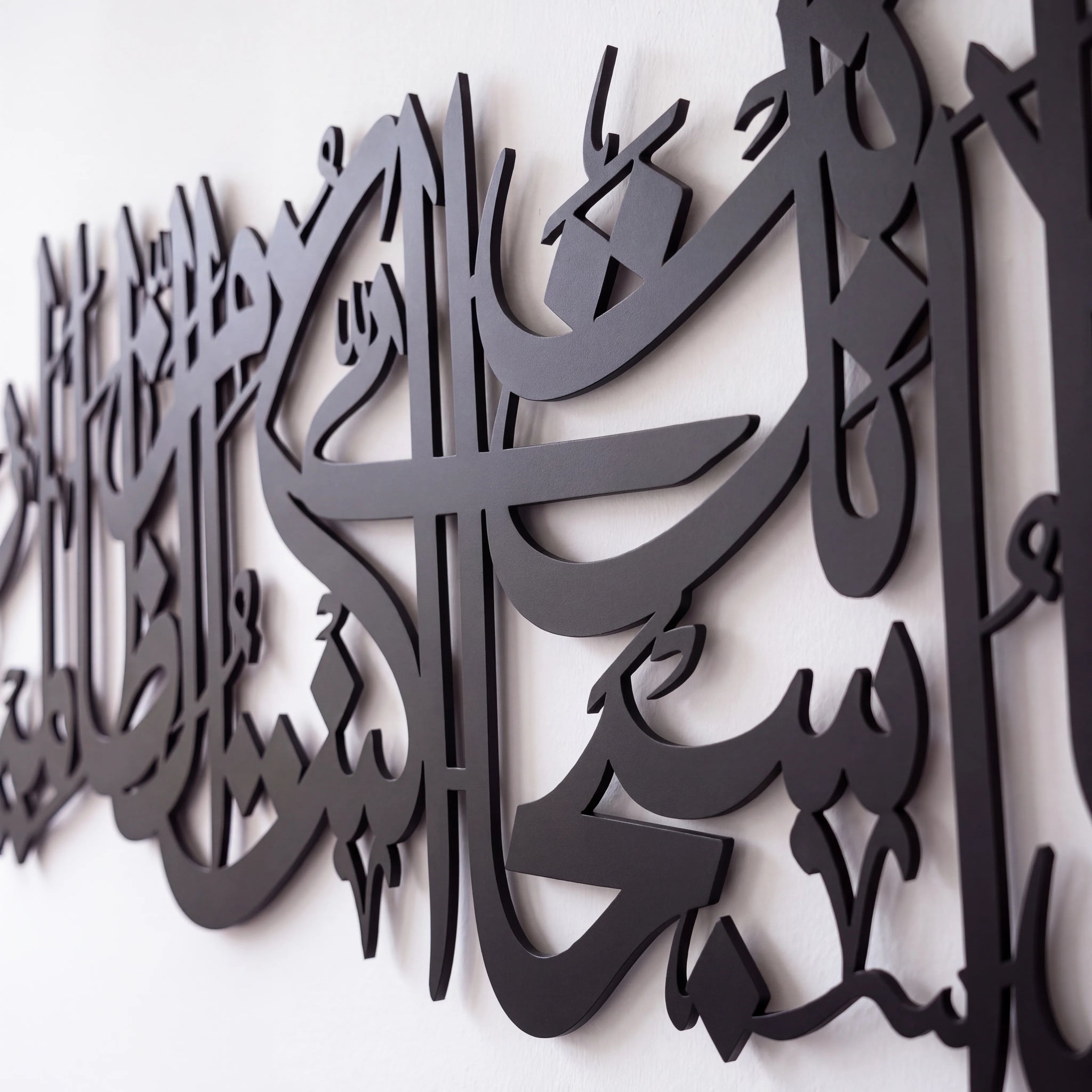 Black metal Islamic wall art featuring elegant Arabic calligraphy, shown mounted on a plain white wall.