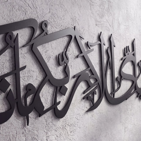 Black metal Islamic wall art featuring Arabic calligraphy mounted on a textured light gray concrete wall.