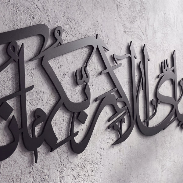 Black metal Islamic wall art featuring Arabic calligraphy mounted on a textured light gray concrete wall.