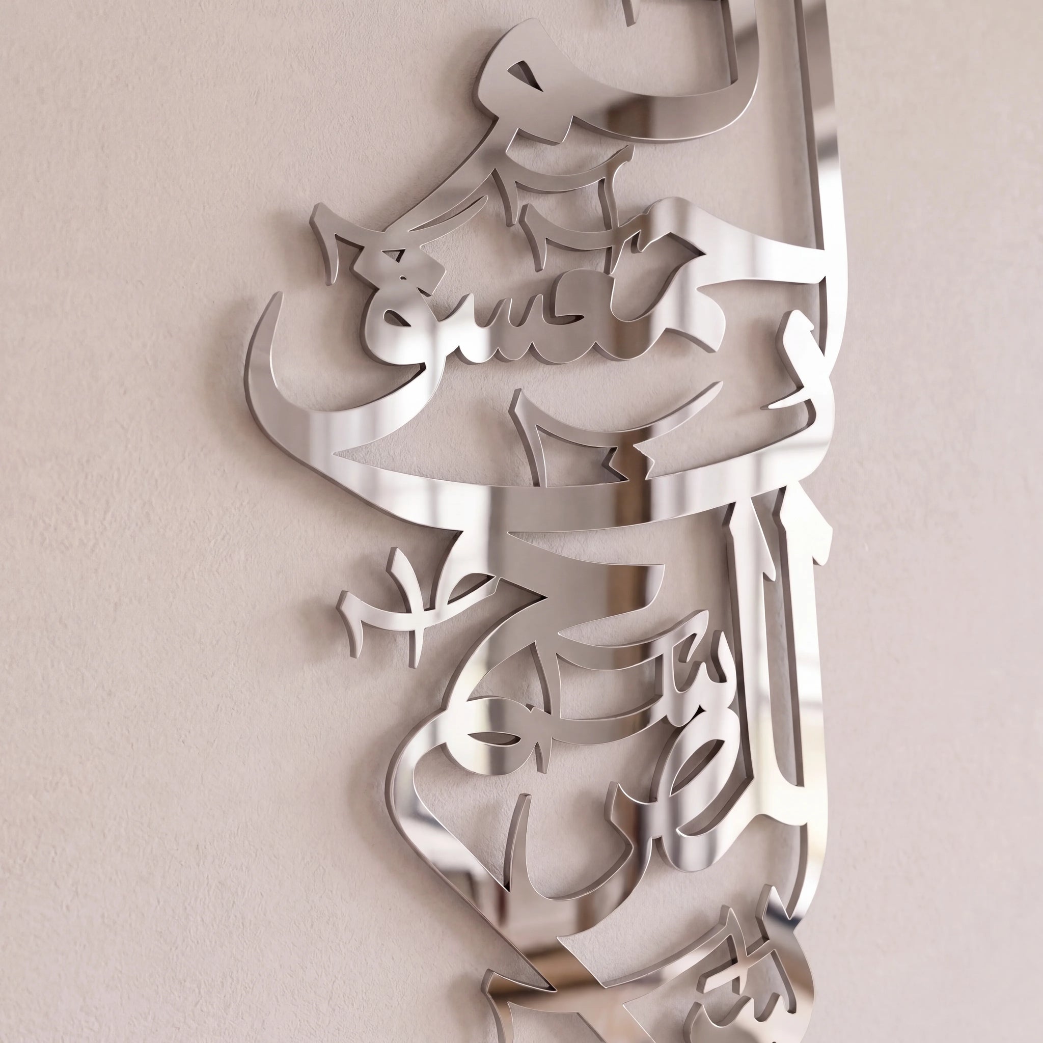 A metallic Arabic calligraphy wall art piece with a polished silver finish, mounted on a light beige textured wall.