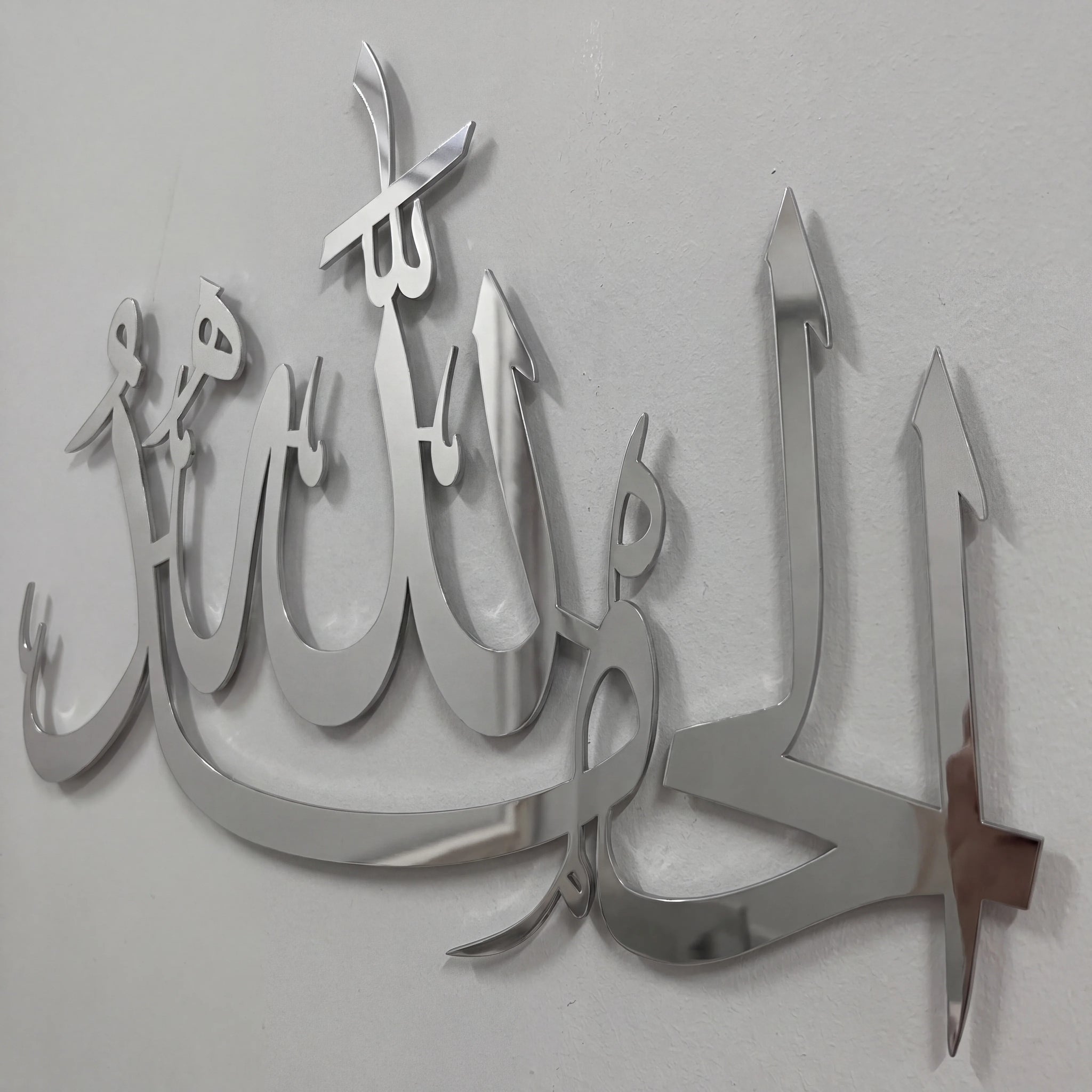 Chrome Silver metallic Arabic calligraphy wall art mounted on a plain light gray wall.
