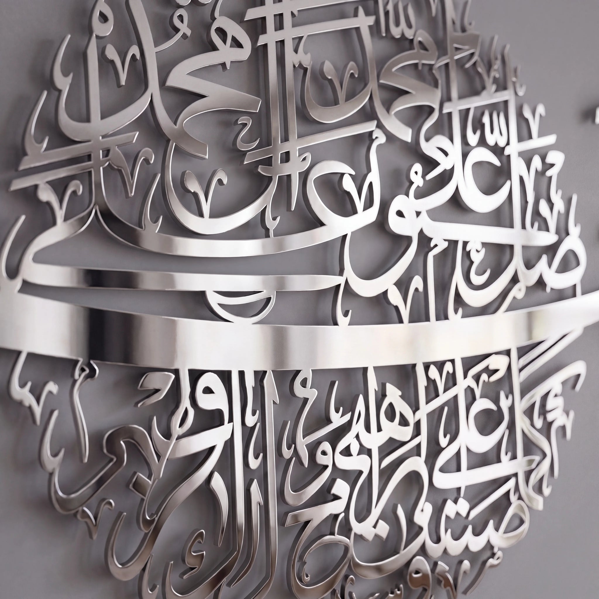 Silver metal Islamic wall art featuring intricate Arabic calligraphy, shown mounted on a gray wall.