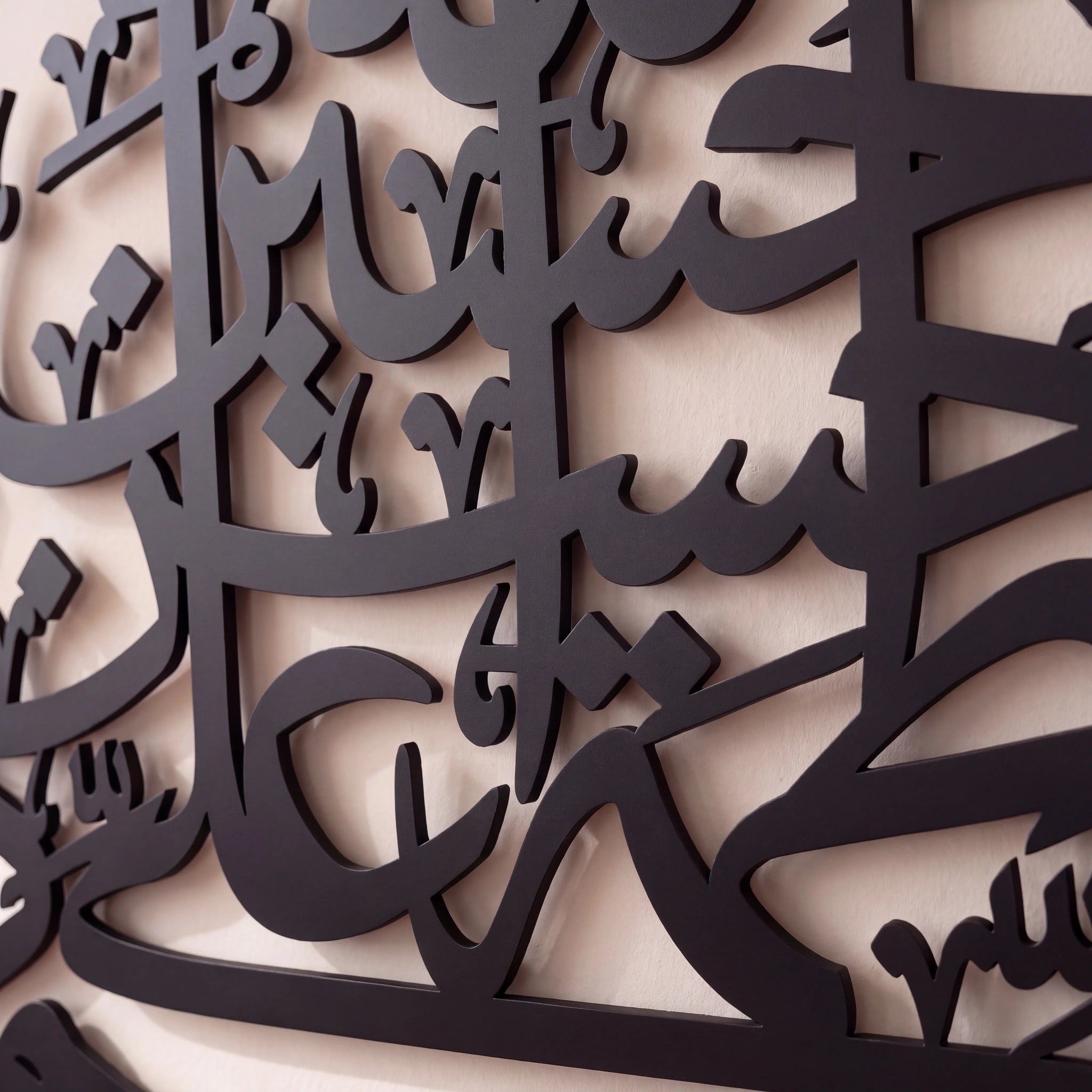Close-up of black metal Islamic wall art featuring elegant Arabic calligraphy layered over a beige wall.