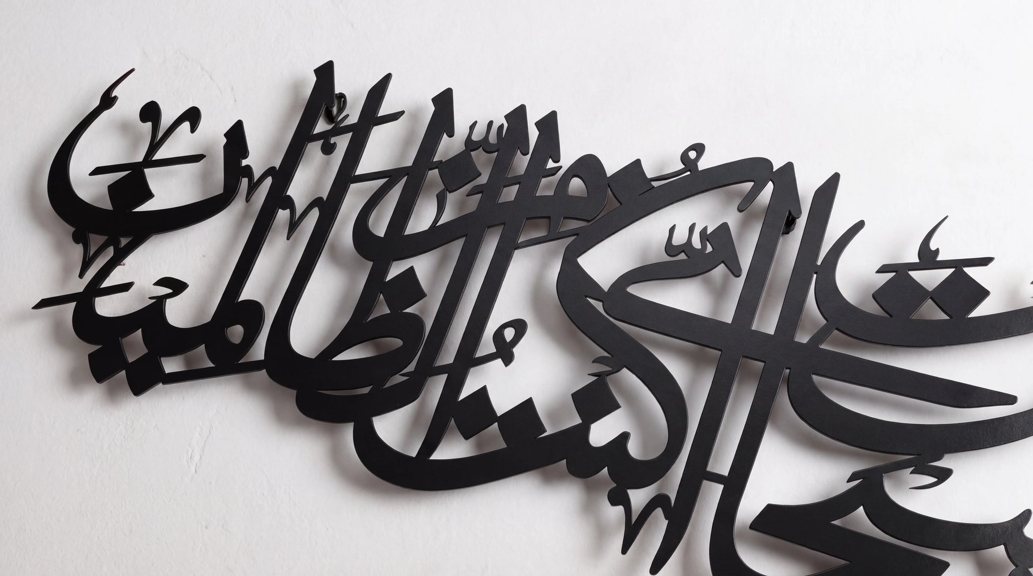Black laser-cut metal Arabic calligraphy wall art mounted on a white wall.