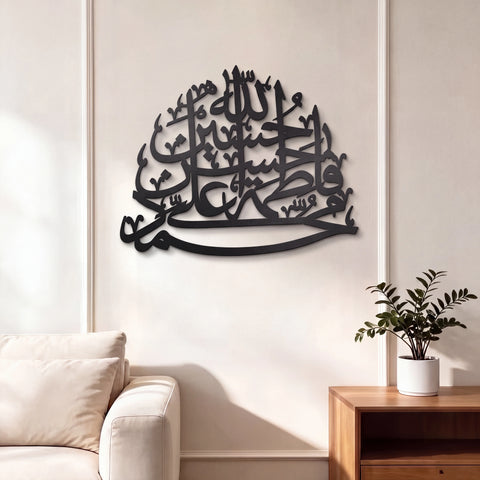 Black Panjtan Pak Metal Islamic wall art featuring Arabic calligraphy, displayed on a cream-colored wall in a modern living room setting with a sofa and wooden side table.