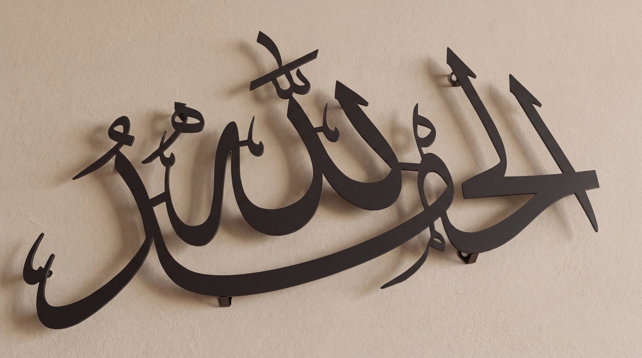 Black metal Islamic wall art featuring elegant Arabic calligraphy, mounted on a plain beige wall.