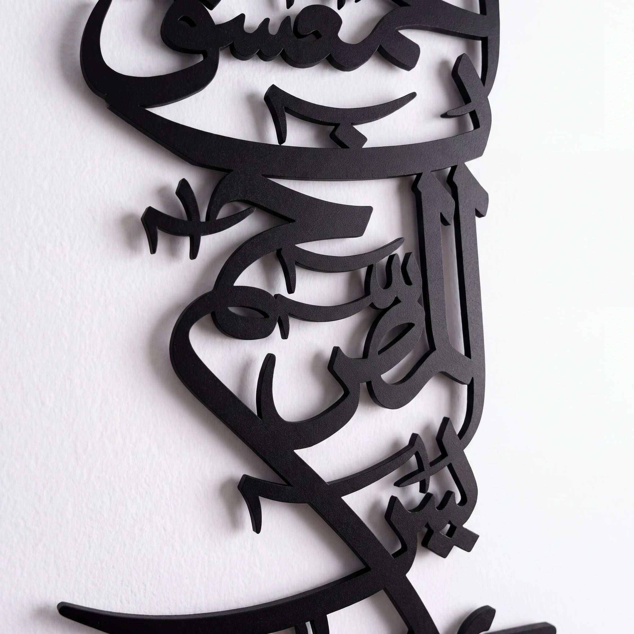 Black metal Islamic calligraphy wall art mounted on a white textured wall.