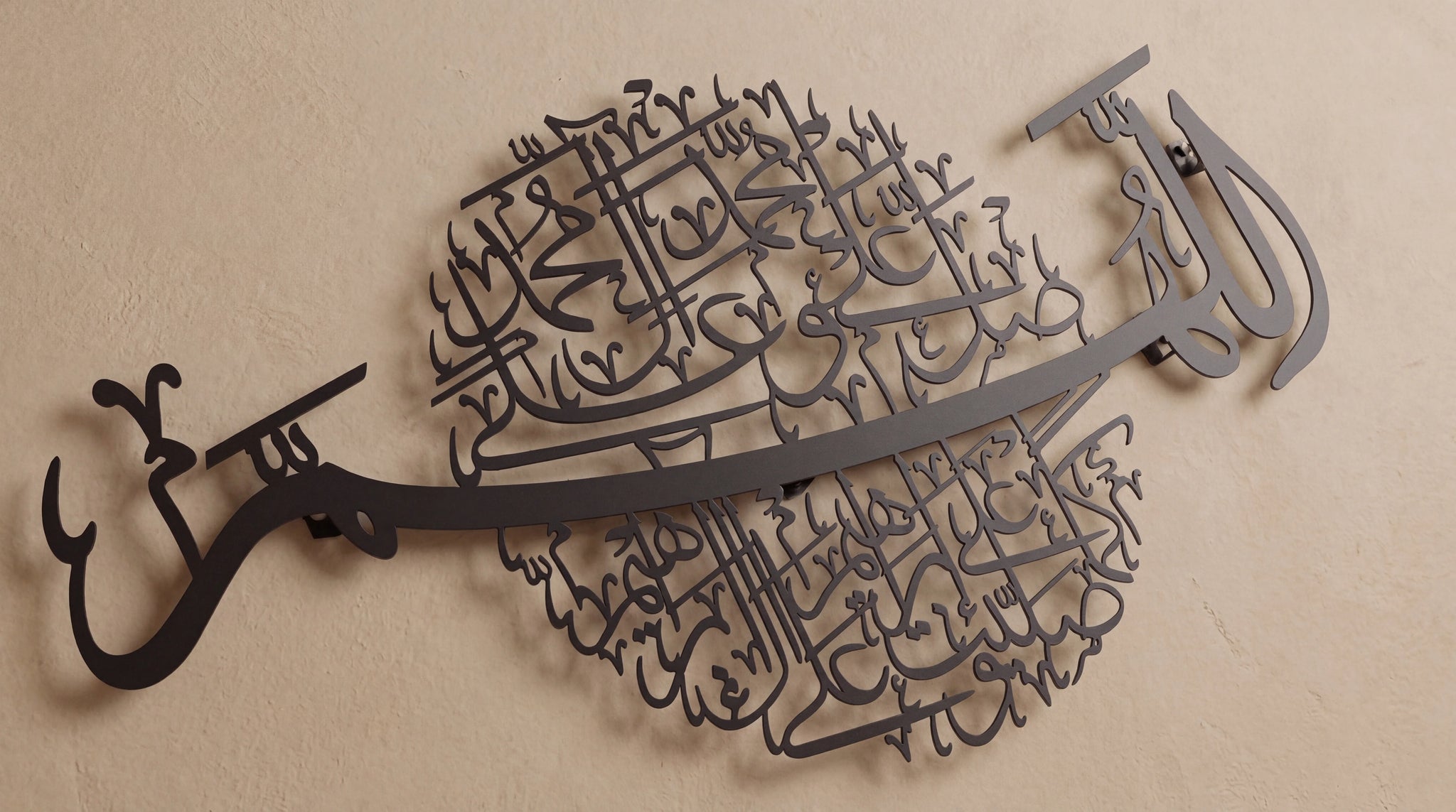 Dark metal Islamic wall art featuring Arabic calligraphy, displayed on a textured beige wall.