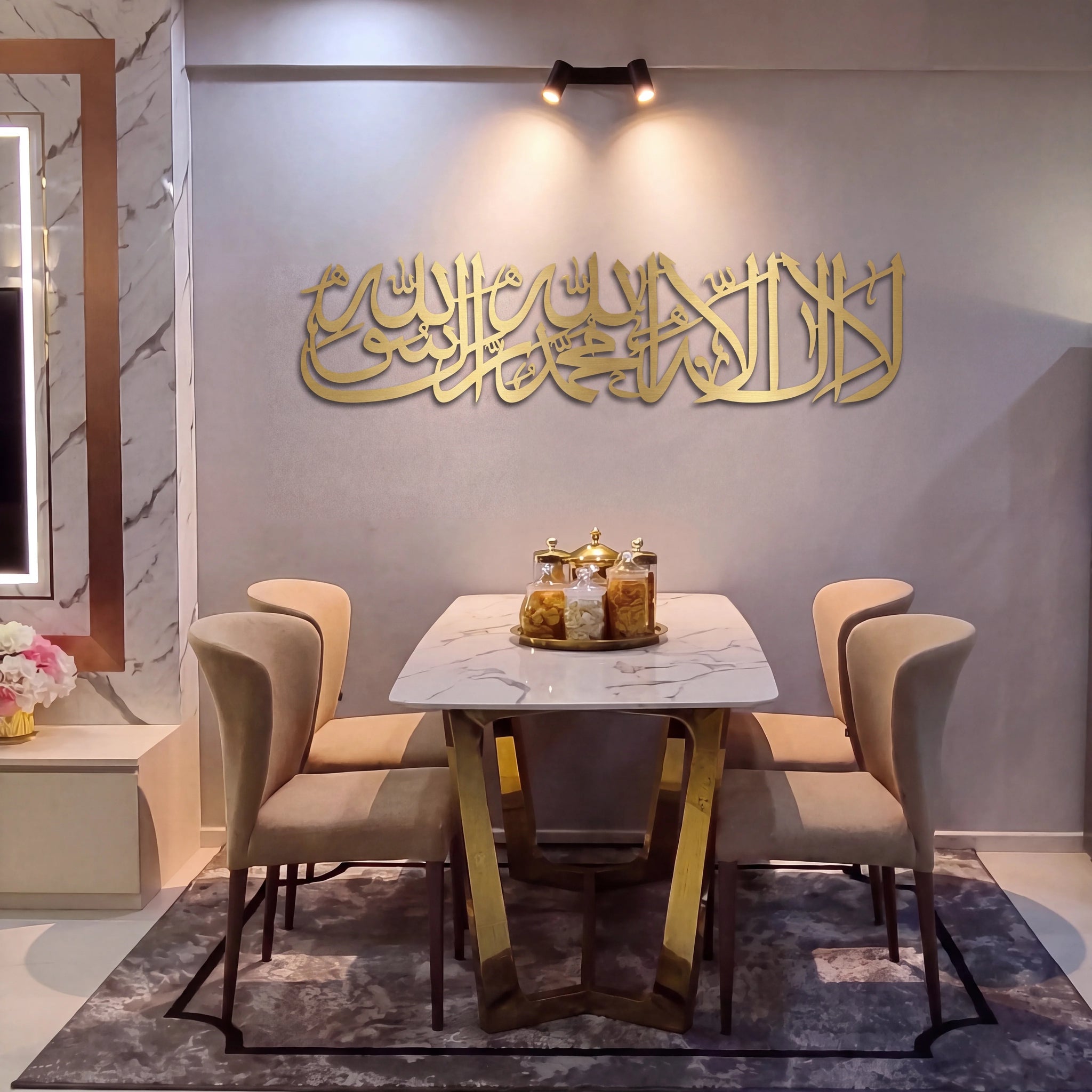 A modern dining room setup featuring a white marble-top table with gold geometric legs, surrounded by four beige upholstered chairs. The table is set on a gray patterned rug against a light gray wall adorned with Mate gold Arabic calligraphy.