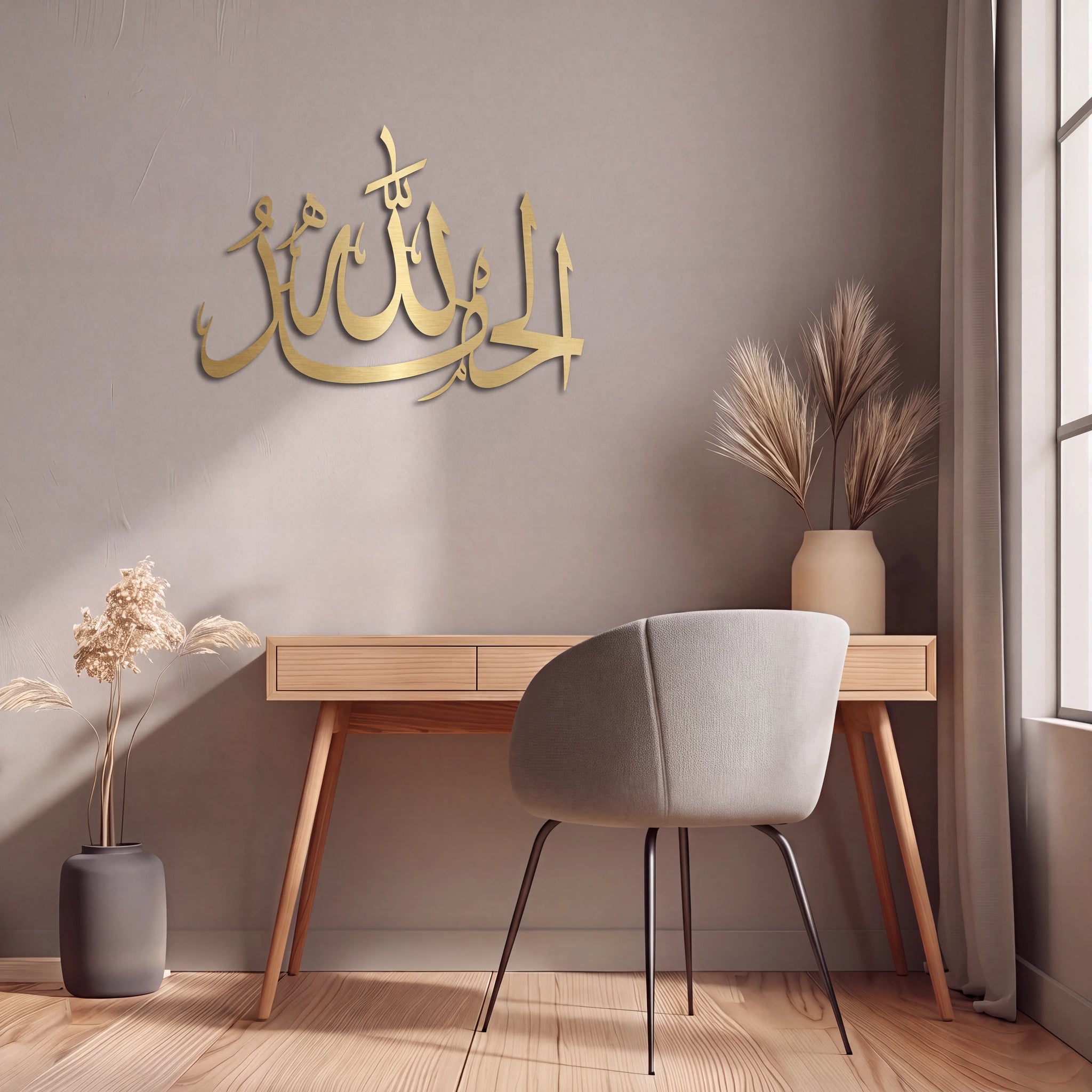 Mate Gold Alhamdulillah Arabic calligraphy wall art mounted on a taupe-gray wall above a light wood desk and gray chair, styled in a modern home office setting with dried plants and a window.