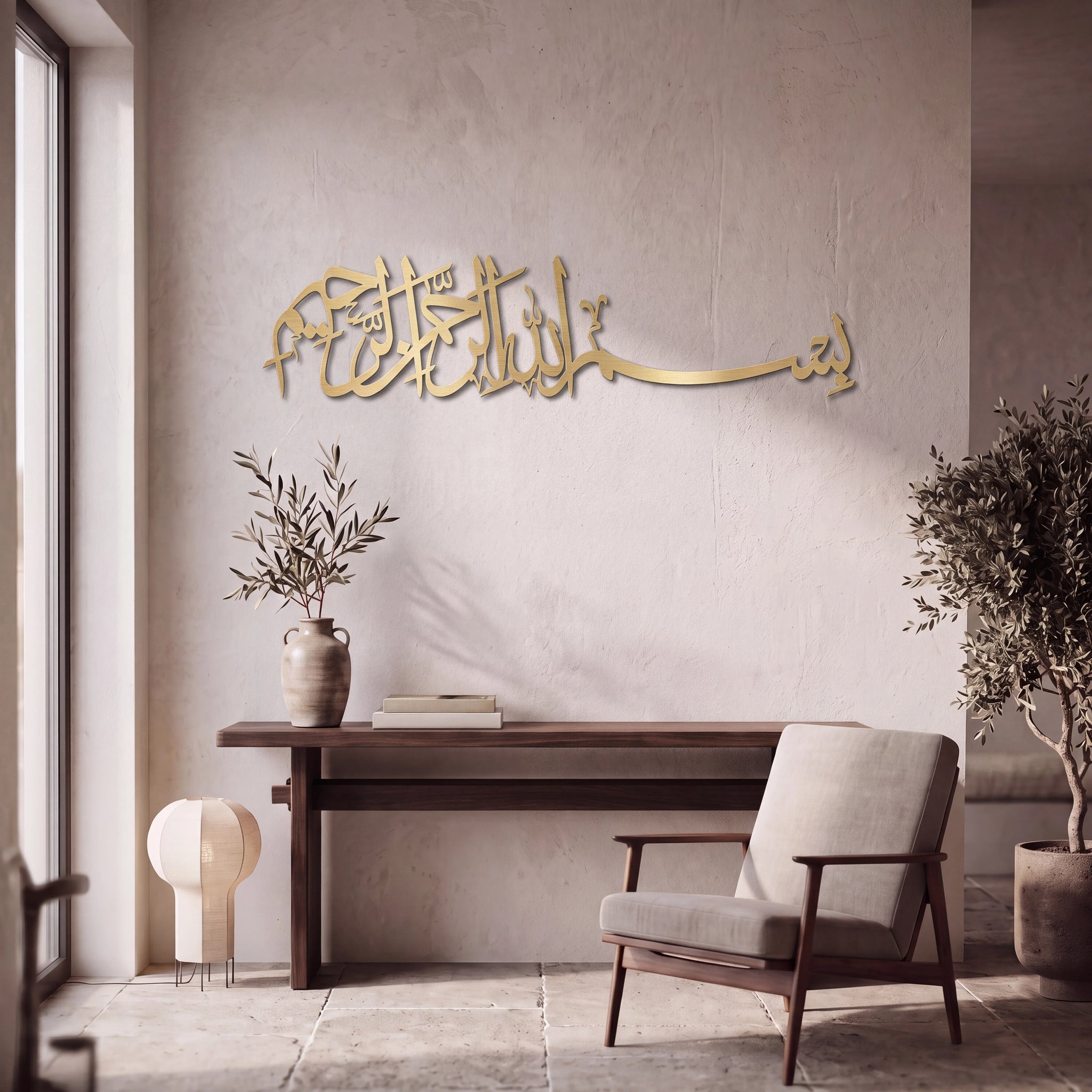 Mate Gold Bismillah Arabic calligraphy wall art mounted on a textured beige wall in a modern, minimalist living room setting with a wooden console table, armchair, and potted plants.