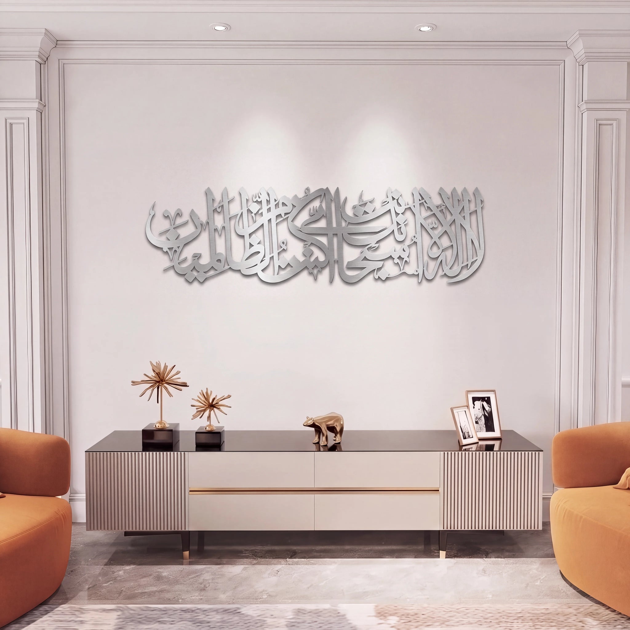 A modern interior scene showing a long mate silver Ayat Kareema Arabic calligraphy wall art piece mounted on a white wall above a low console table, with orange chairs at the sides and decorative objects on the table.