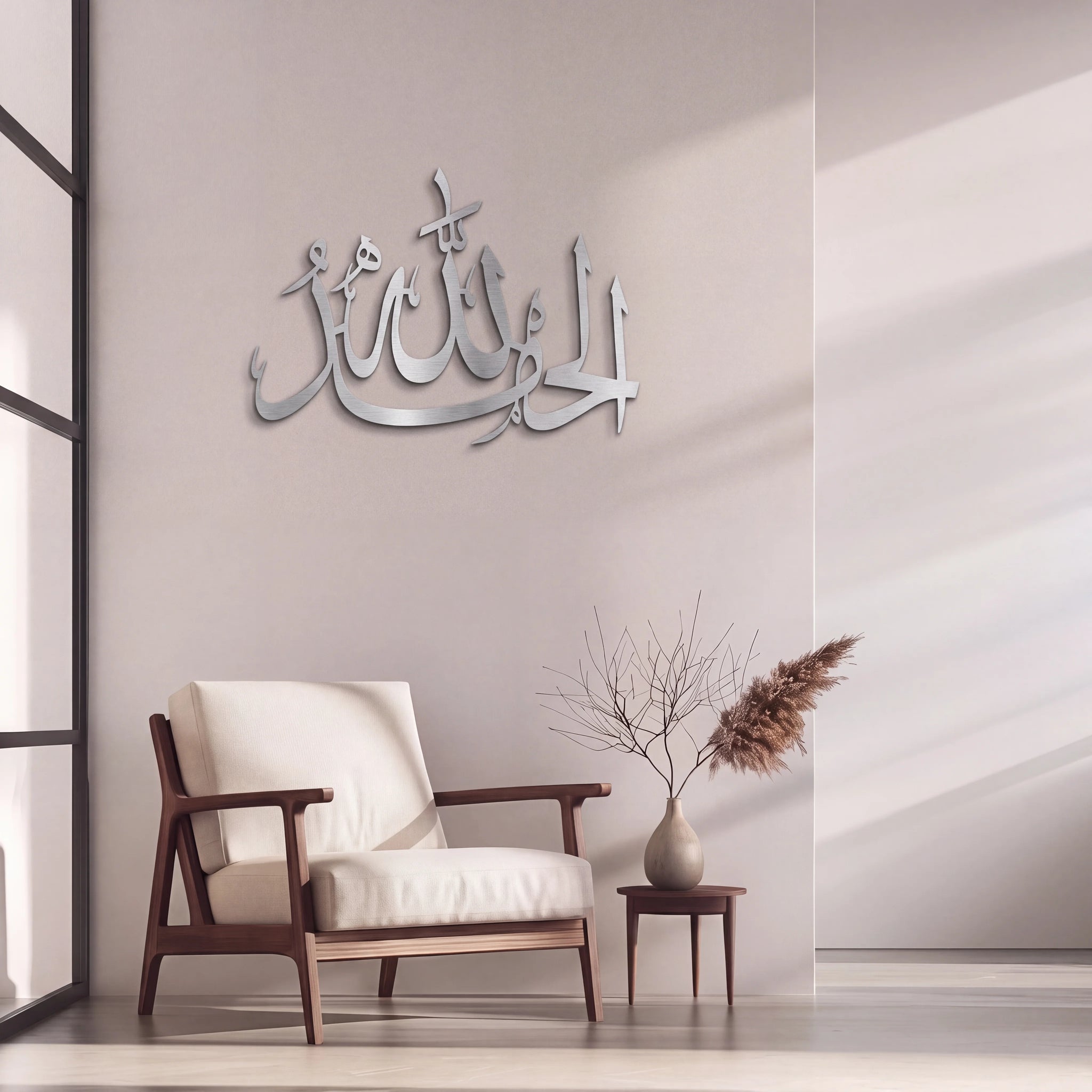 Mate Silver Alhamdulillah Arabic calligraphy wall art mounted on a light beige wall in a modern living room setting, featuring a wooden armchair with white cushions, a small side table with a vase of dried branches, and pampas grass.