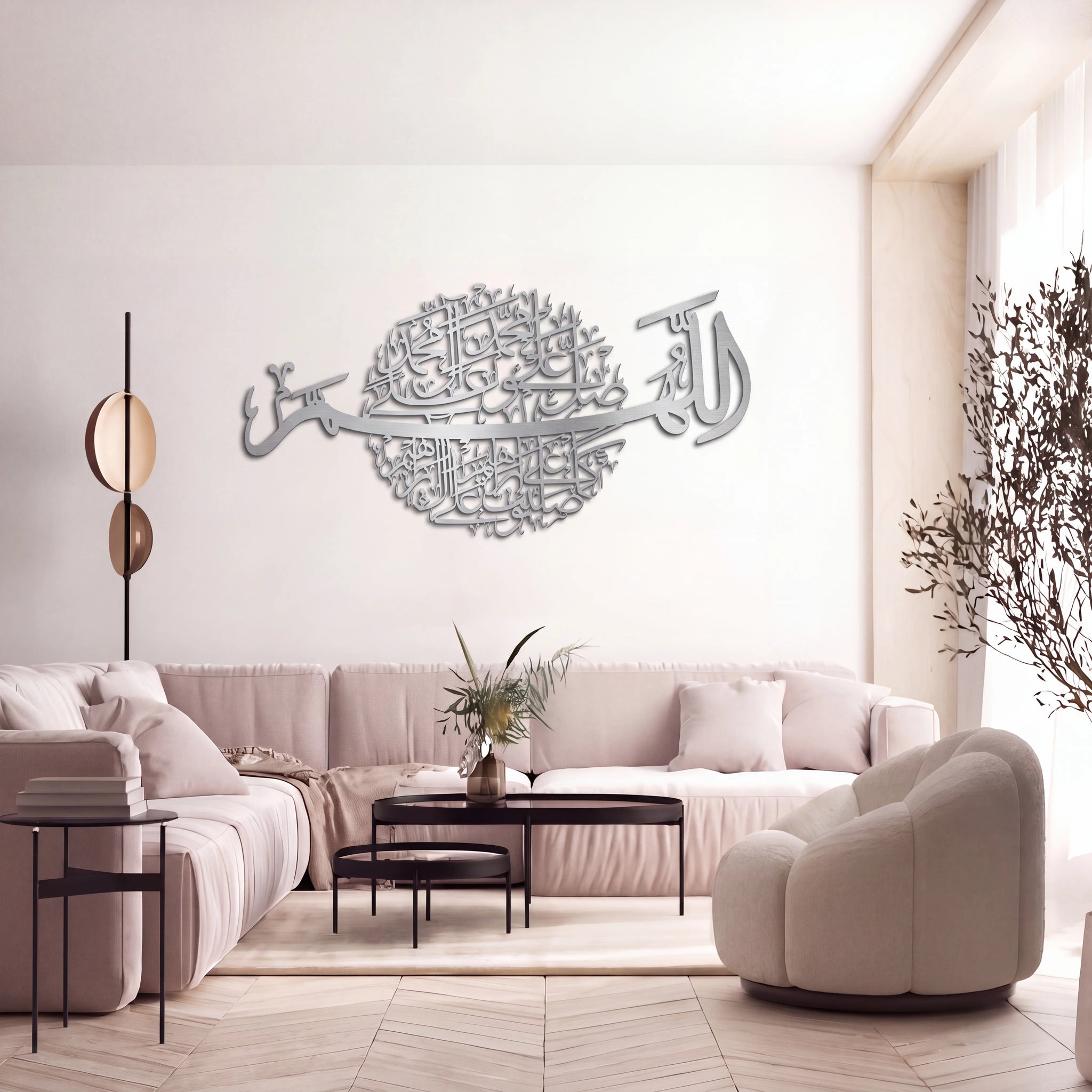A modern living room with Darood Shareef large Mate silver Arabic calligraphy wall art piece mounted on a white wall above a light pink sofa. The room features a beige armchair, minimalist black tables, a floor lamp, and a light wood herringbone floor.