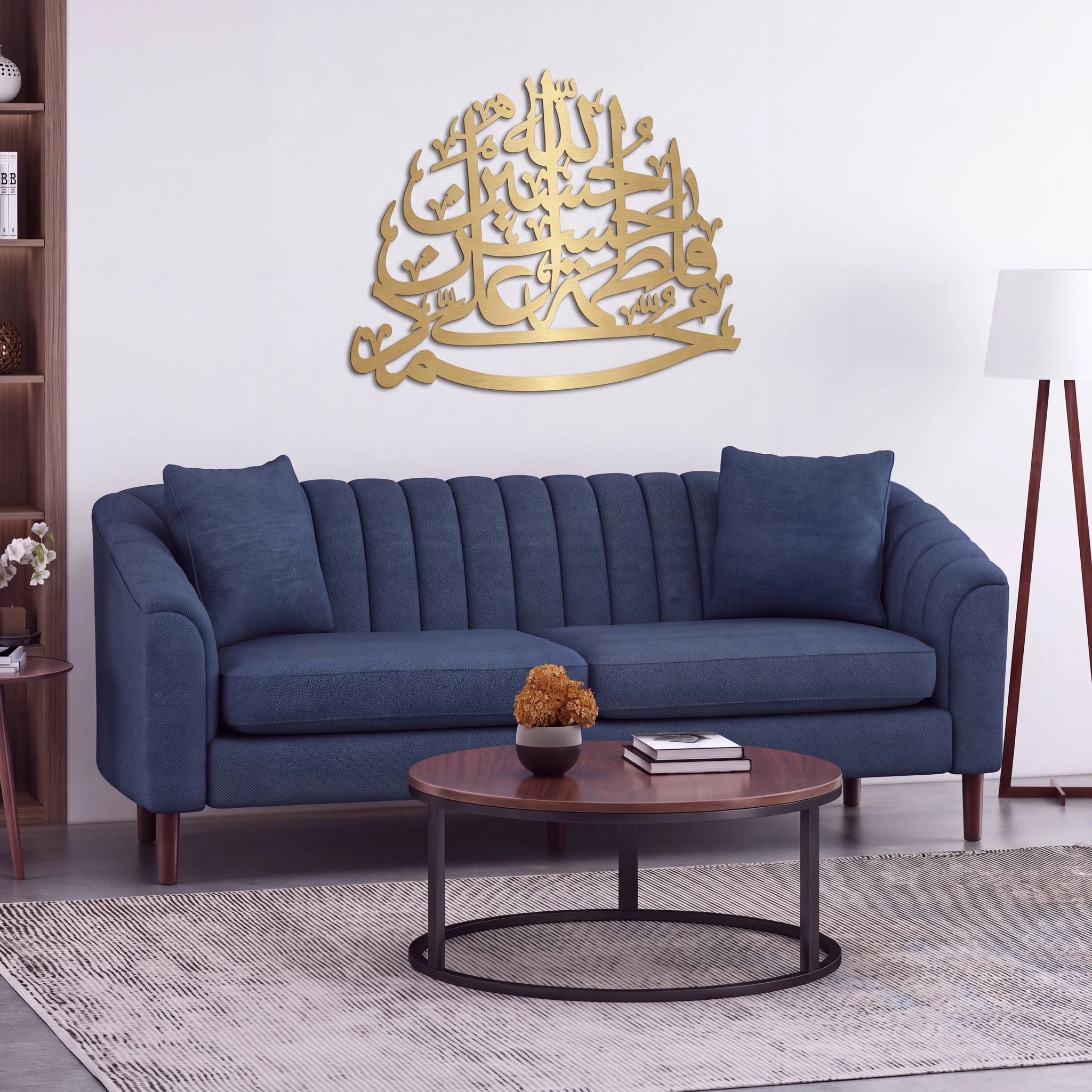 A modern living room setting featuring a navy blue velvet sofa with a large, metallic Mate gold Panjtan Pak Arabic calligraphy wall art piece mounted on the wall above it. In front of the sofa is a round wooden coffee table with a black metal base, set on a textured gray rug.