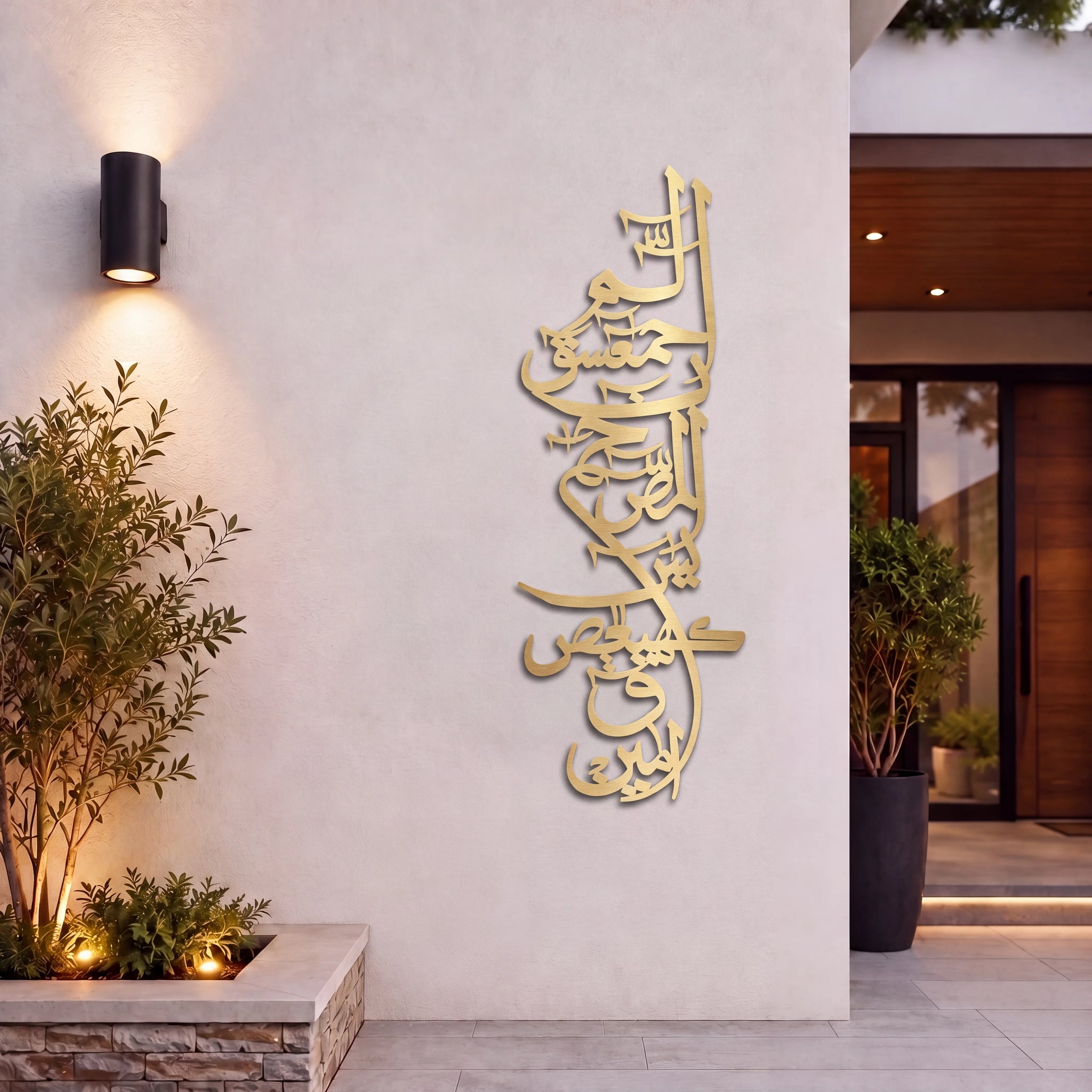 Mate Gold Lohe Qurani Arabic calligraphy wall art mounted on a beige stucco wall in a modern outdoor entryway, with a black wall sconce, potted plants, and warm ambient lighting.