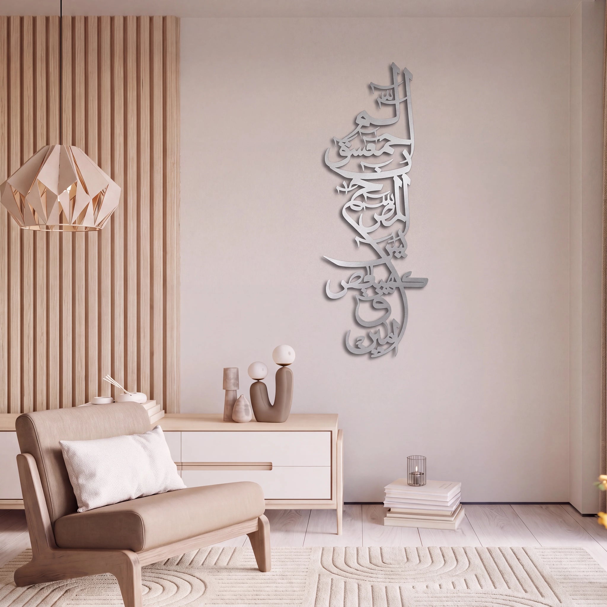 A modern living room setting featuring a tall, vertical Arabic calligraphy wall art piece in a Mate silver finish mounted on a beige wall. The room includes a light wood slatted accent wall, a geometric pendant light, a beige armchair with a white pillow, a light wood side table with decorative vases, and a textured cream rug.
