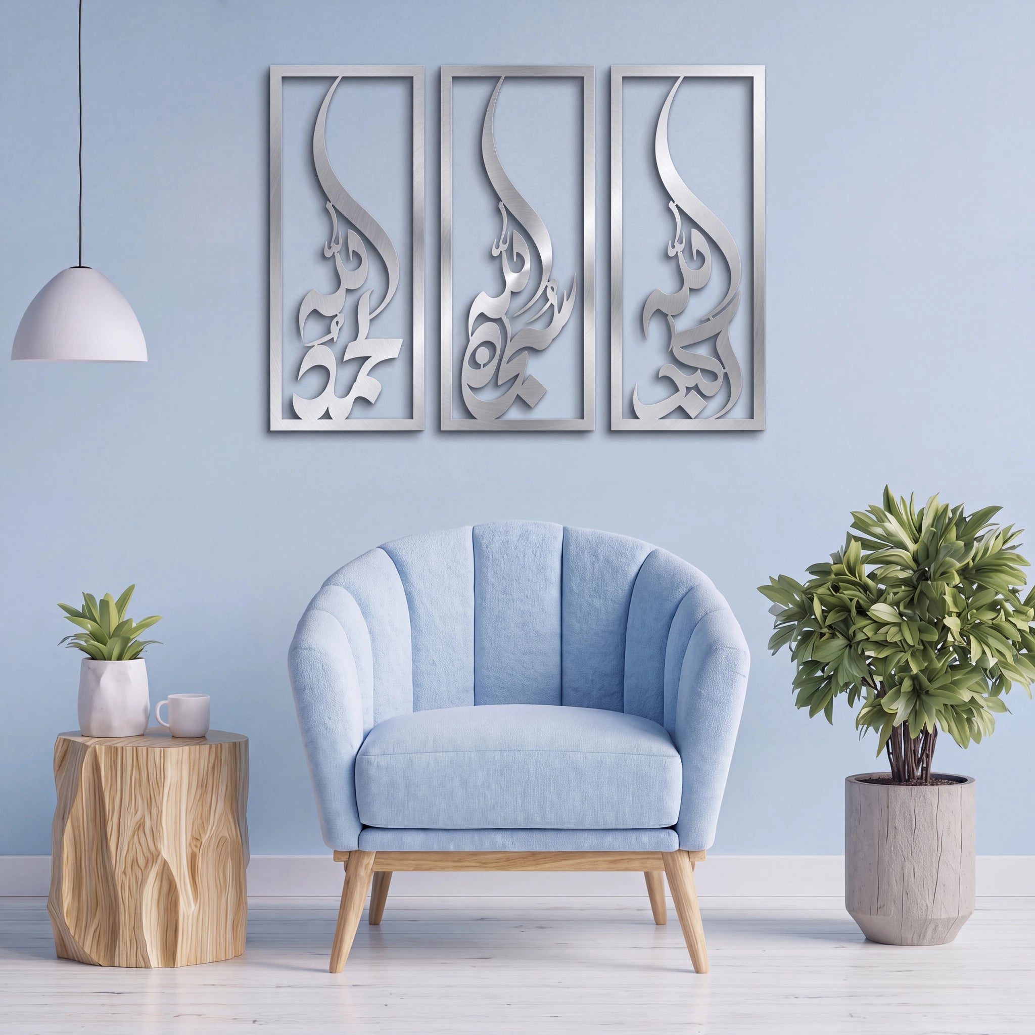 A light blue upholstered accent chair with wooden legs is centered in a styled room against a pale blue wall, with three vertical Subhanallah Alhamdulillah Allah Ho Akbar Mate Silver wall art panels above it, a white pendant light on the left, a small wood-side table with a plant on the left, and a larger potted plant on the right.
