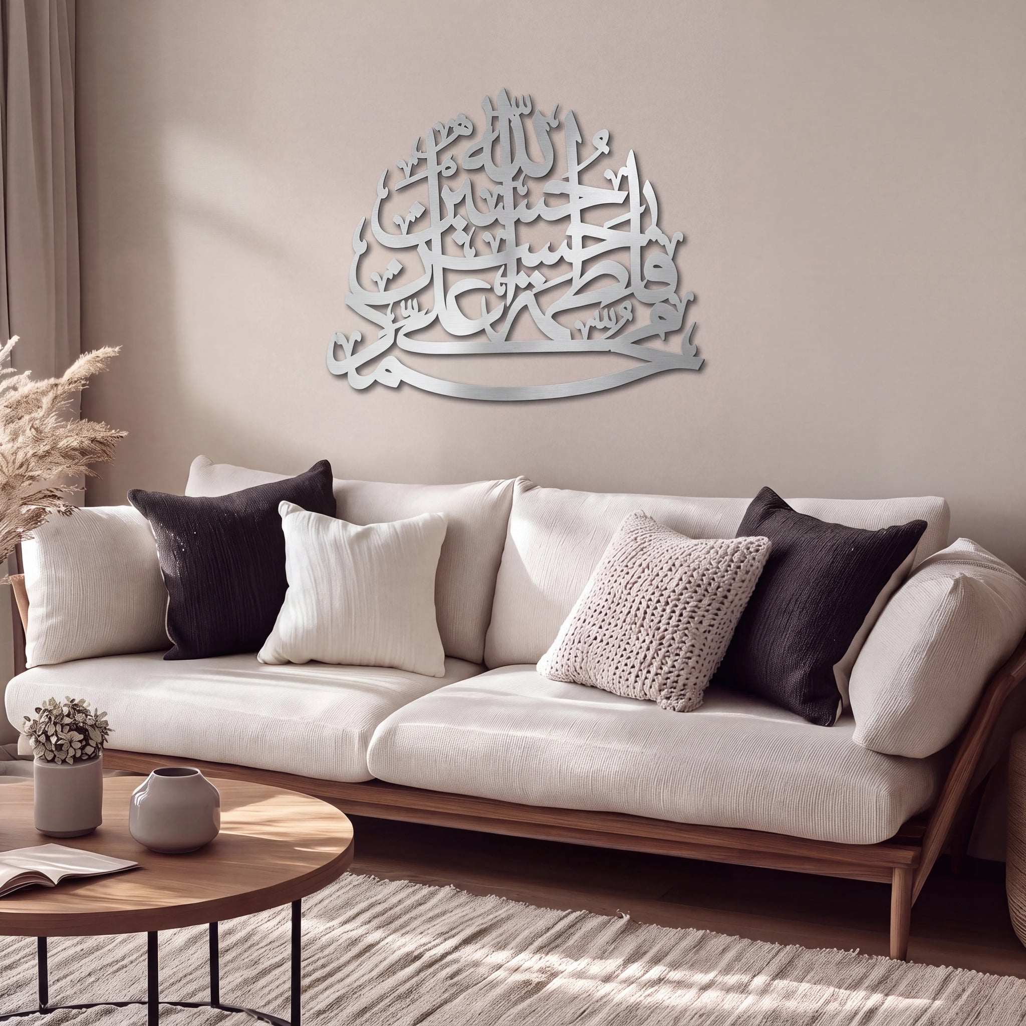 A modern living room scene featuring a white upholstered wooden sofa with black and cream throw pillows, decorated with a large mate silver Panjtan Pak Arabic calligraphy wall art piece on the beige wall above it.